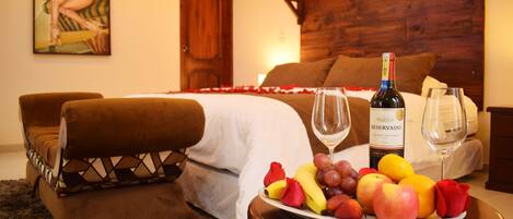 A bedroom with a wooden headboard, a bed with white linens, a bedside table with a bottle of wine and glasses, and a fruit platter on the table.