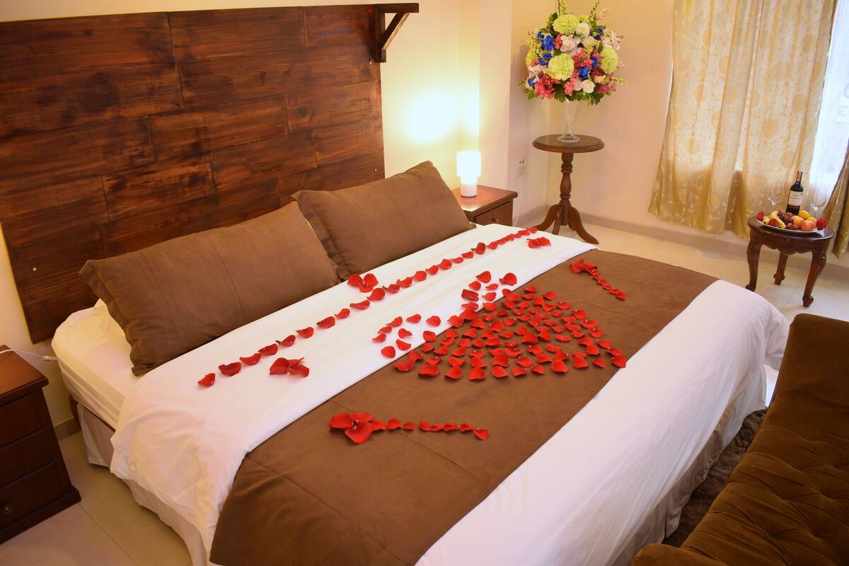 A neatly made bed with a brown headboard, a bedside table with a lamp, and a small table with a vase of flowers.