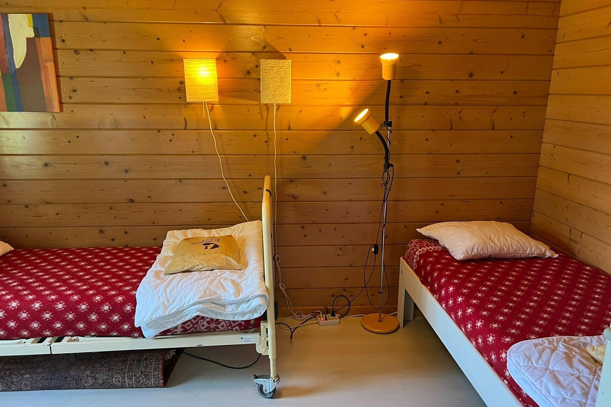 A wooden cabin room with two beds, each with a red patterned bedspread and white pillows. There are two wall-mounted lamps providing light.