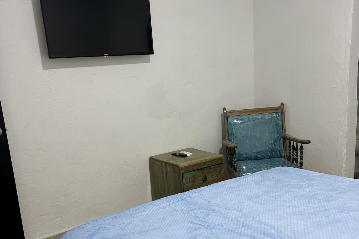 A bedroom with a bed, a nightstand, a chair, a television on the wall, and a small wooden cabinet.