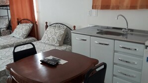 Une cuisine compacte avec un coin repas, un lit avec une literie à motifs et une fenêtre agrémentée de rideaux orange.