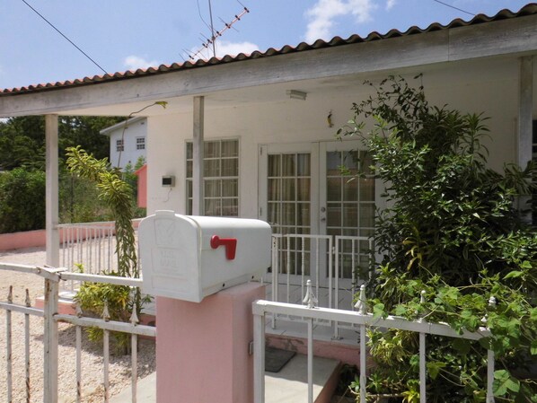 A white mailbox with a red flag, attached to a pink post, in front of a white house with a tiled roof and a white railing. - Curacao Vacation Homes (Willemstad)