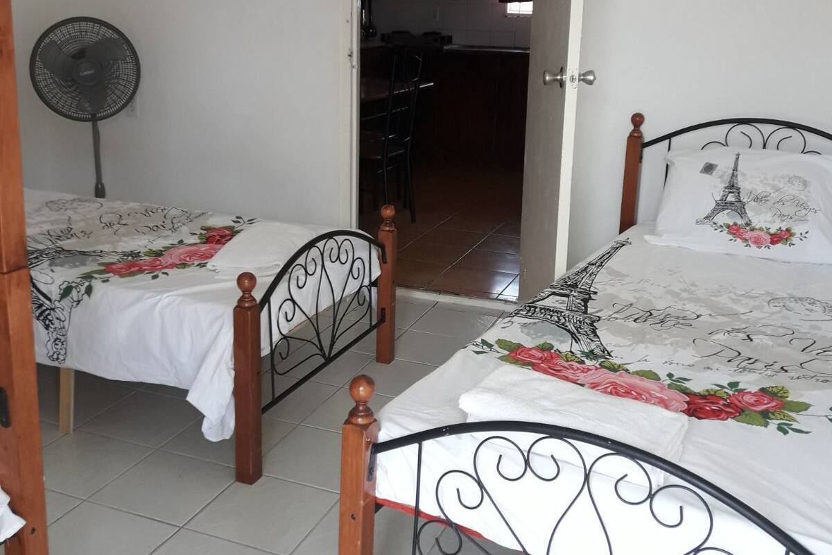 A bedroom with two beds, a fan, and a view of a kitchen and dining area through an open door.