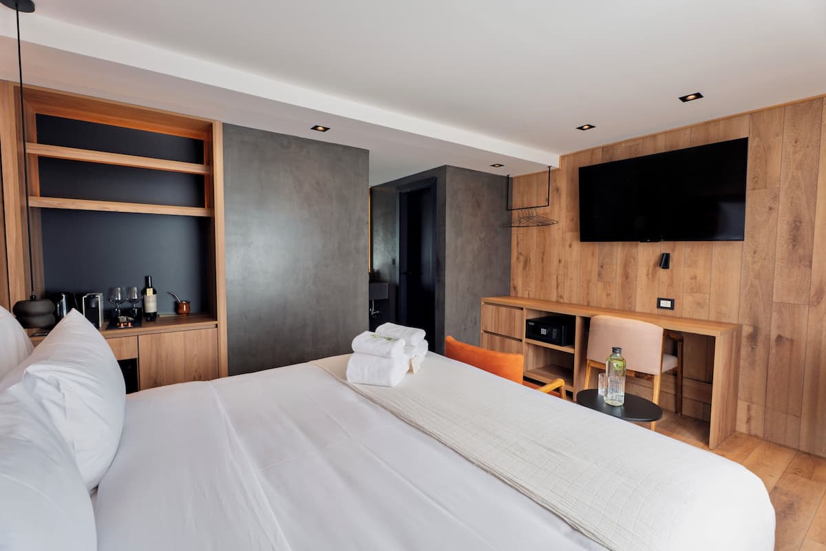 A modern hotel room with a large bed, a flat-screen TV mounted on the wall, a wooden desk, and a seating area with a chair.