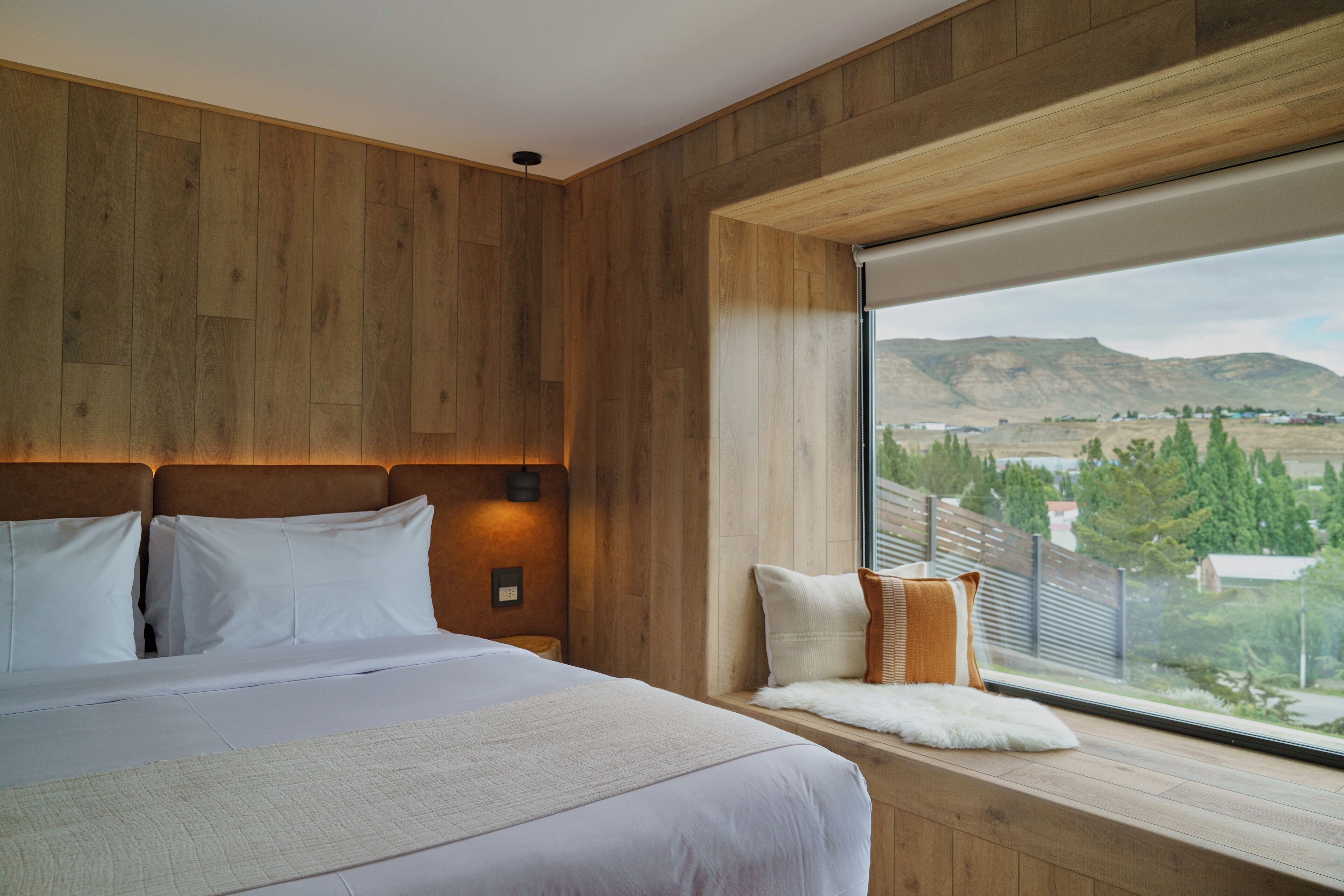 Un dormitorio con una cama grande, paredes de madera, un ventanal con vistas al campo y un pequeño banco con cojines.