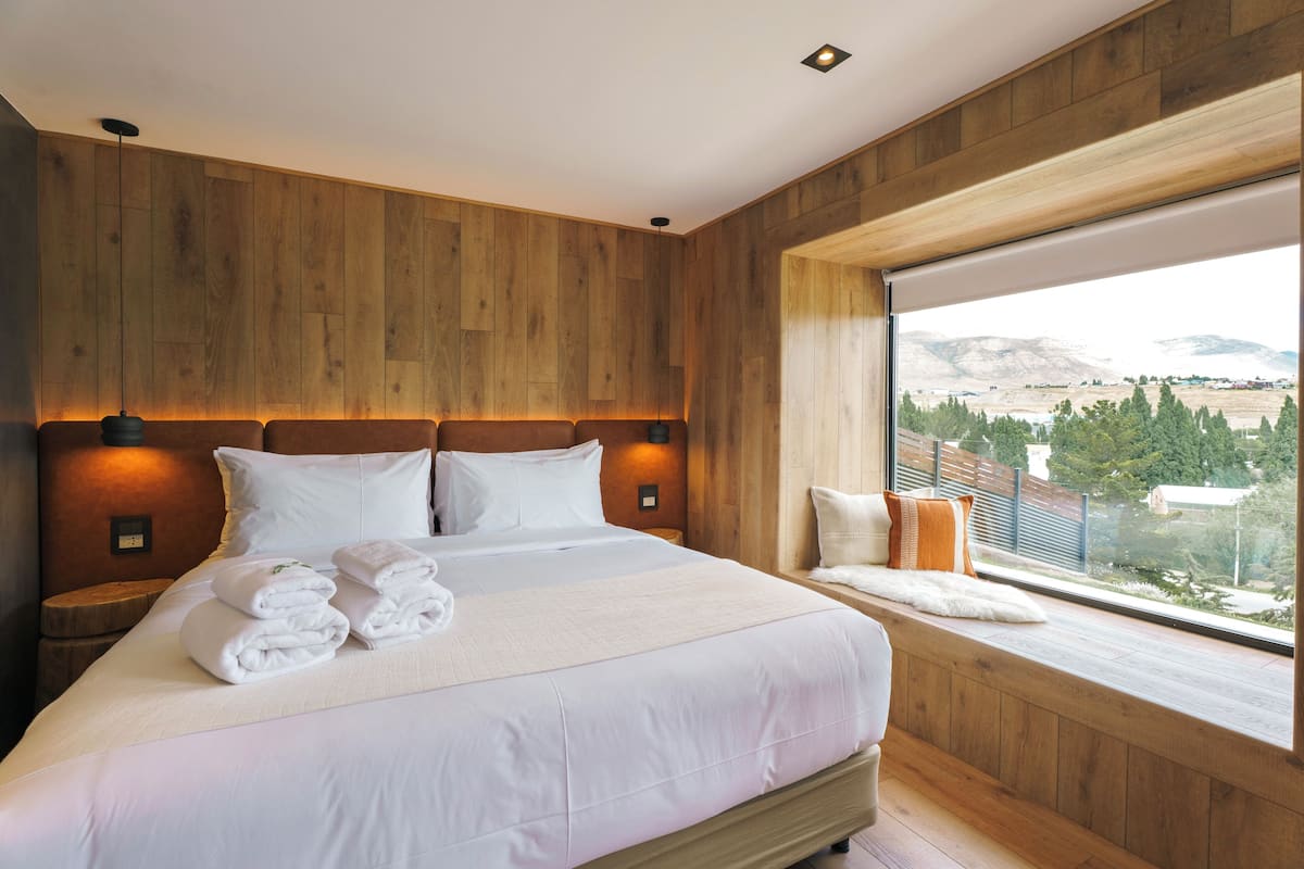 A modern hotel room with a large bed, wooden panel walls, a large window with a view, and a small seating area by the window.