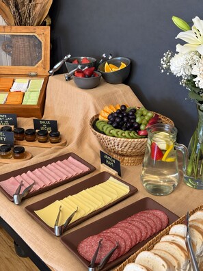 Una mesa de buffet con diversos alimentos, como carnes frías, quesos, frutas y una jarras de bebidas.