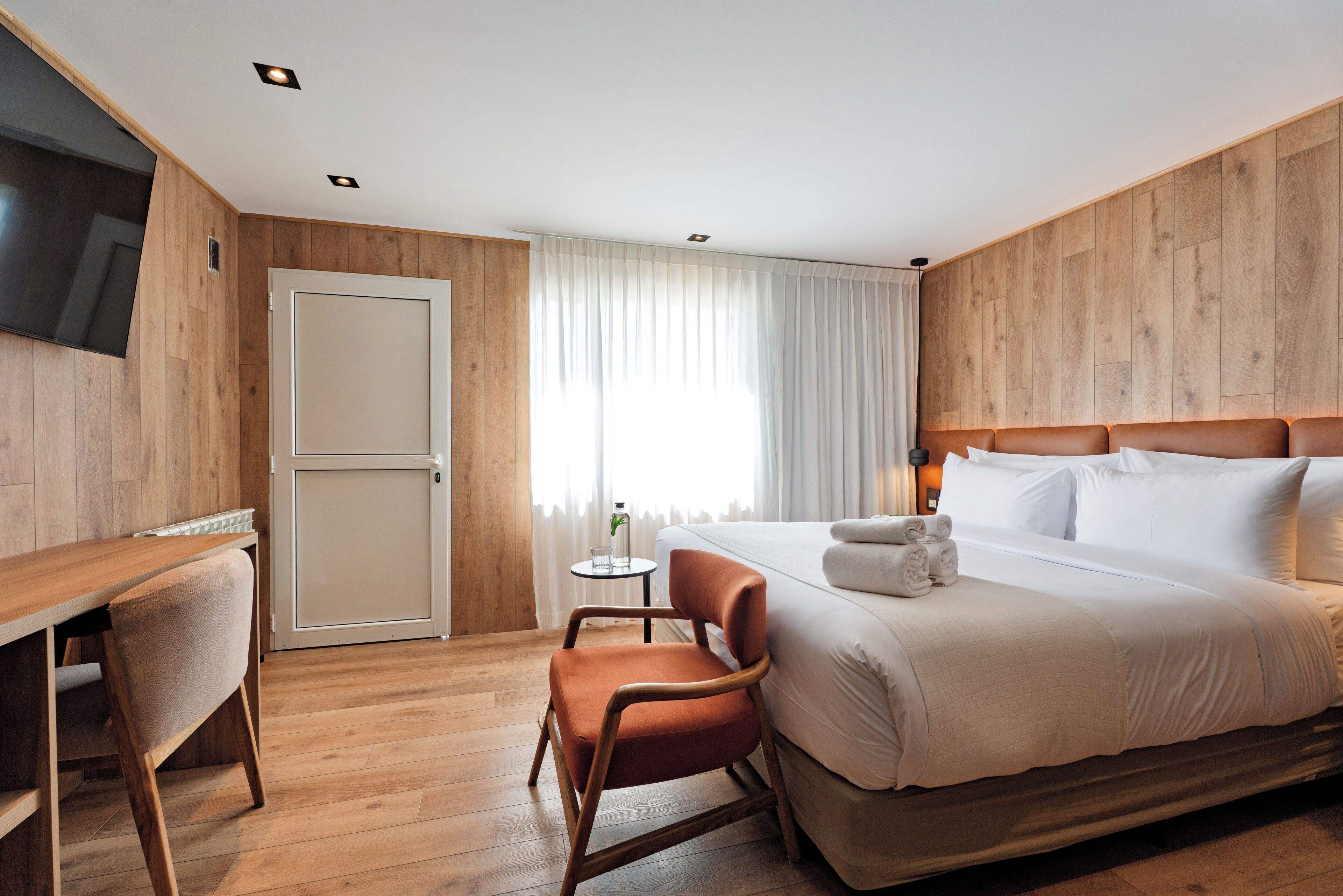 A hotel room with a wooden floor, a bed with white linens, a wooden desk with a chair, a small table with a glass and a plant, and a door.