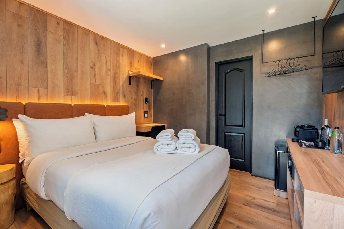 A modern hotel room with a wooden headboard, a neatly made bed with white linens, a small bedside table, a built-in desk with a coffee machine, and a dark door.