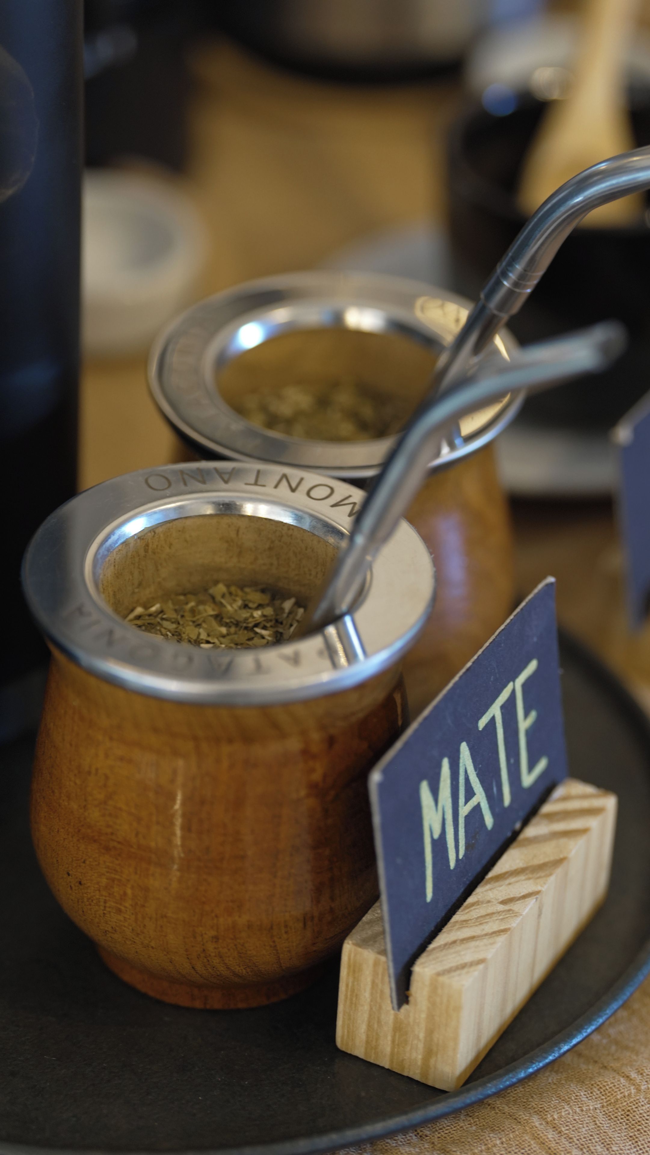 Två mate-krus med lock av metall och en sked inuti, placerade på en mörk tallrik med ett underlägg av trä märkt med texten ”MATE”.