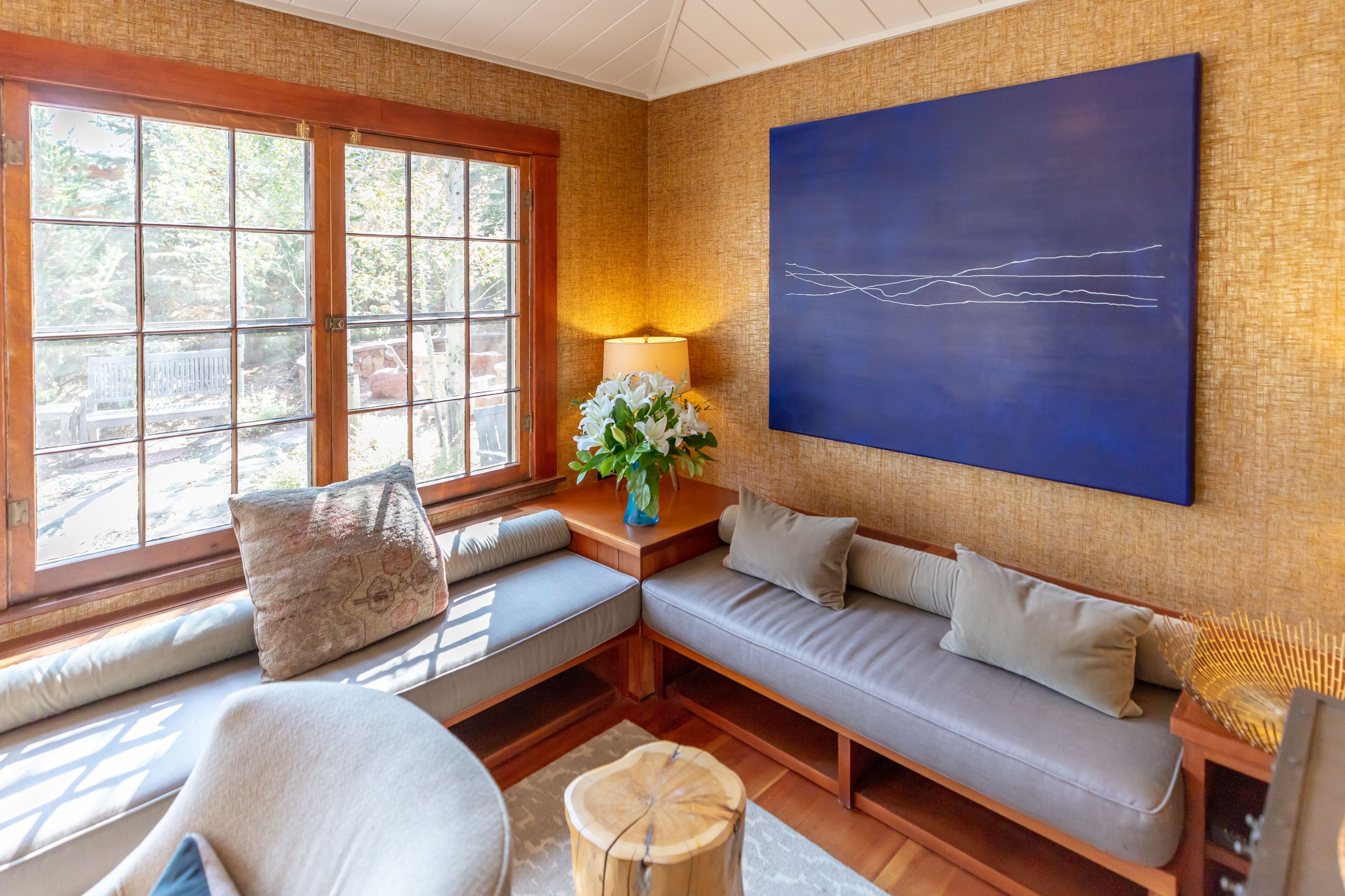 A cozy room with a large window, a sofa with cushions, a lamp, and a blue abstract painting on the wall.