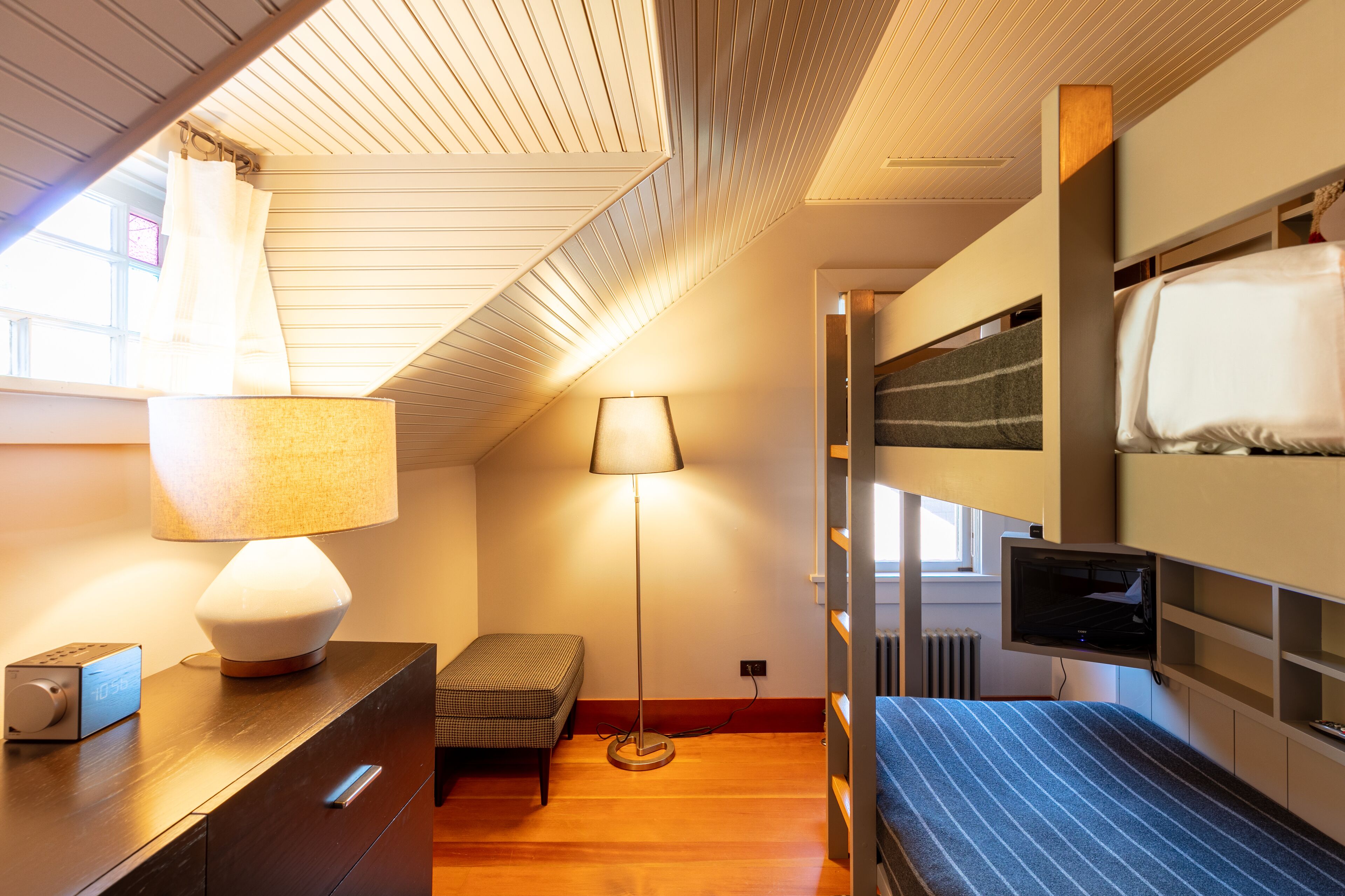 Ein kompakter, gut beleuchteter Raum mit einem Etagenbett, einem Nachttisch mit Lampe, einem kleinen Schreibtisch und einem an der Wand montierten Fernseher.