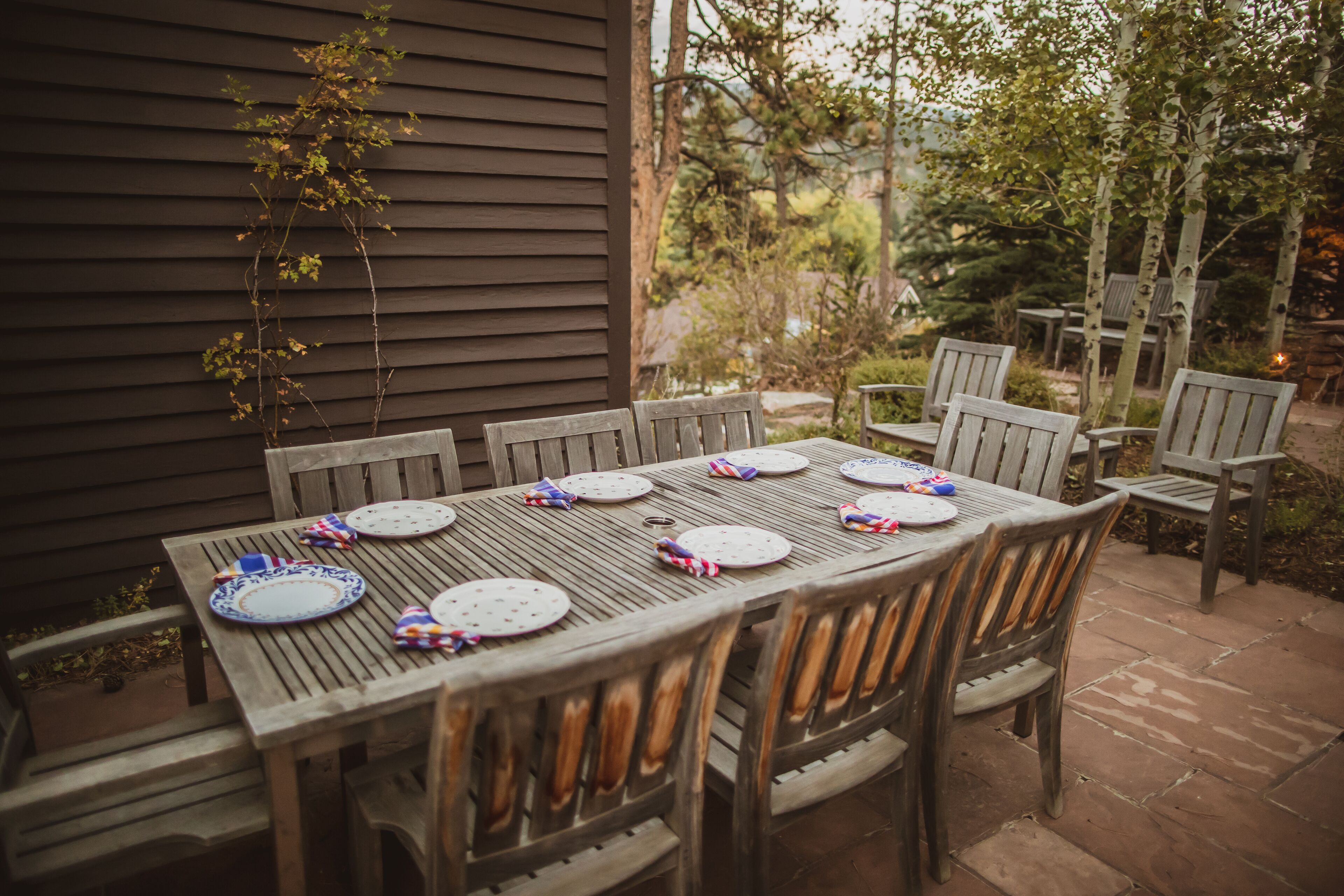 An outdoor dining area with a wooden table and chairs, set against a house with a dark siding exterior.