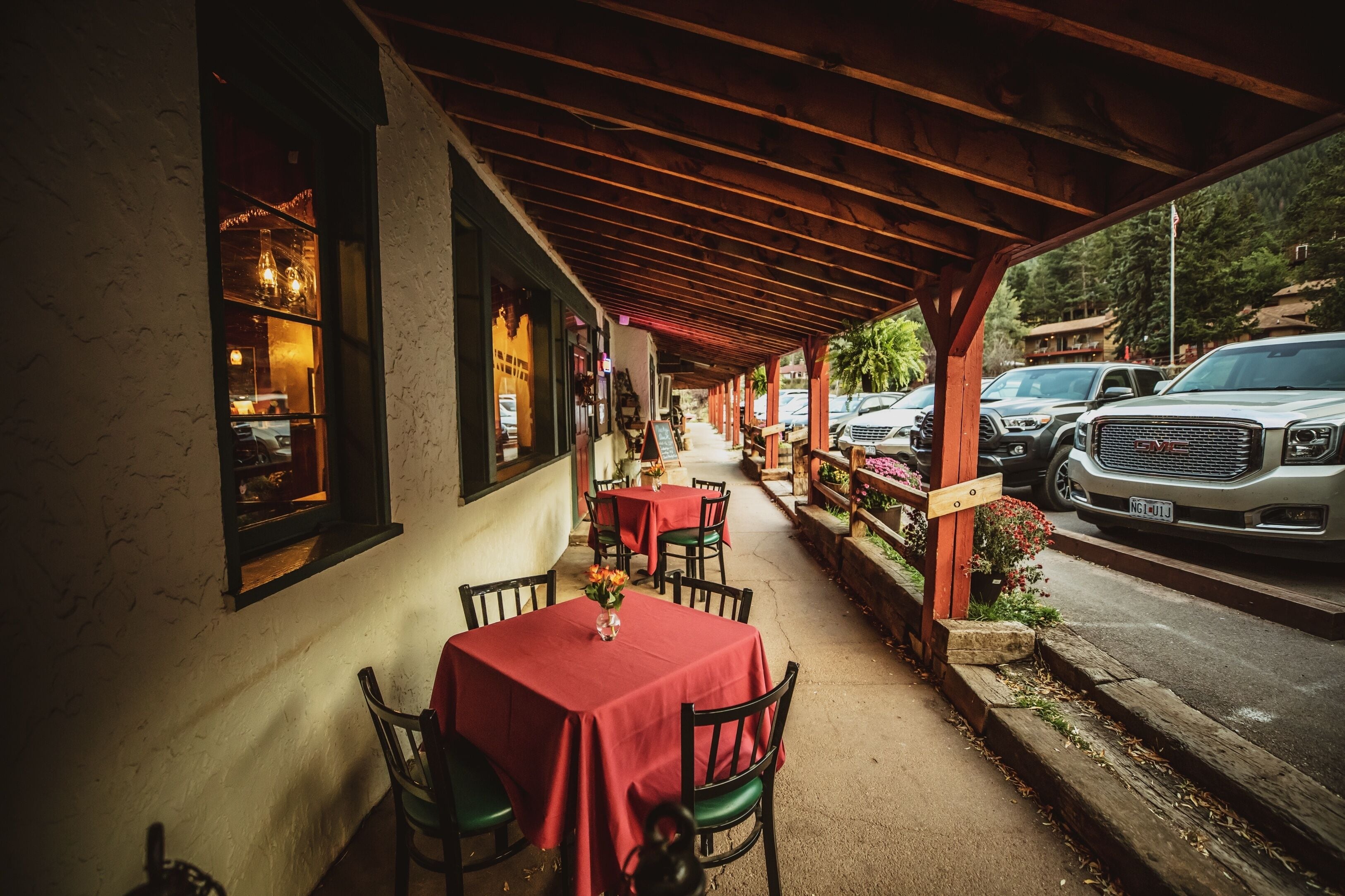 An outdoor dining area with tables and chairs, a covered walkway, and parked cars.