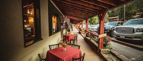 An outdoor dining area with tables and chairs, a covered walkway, and parked cars.