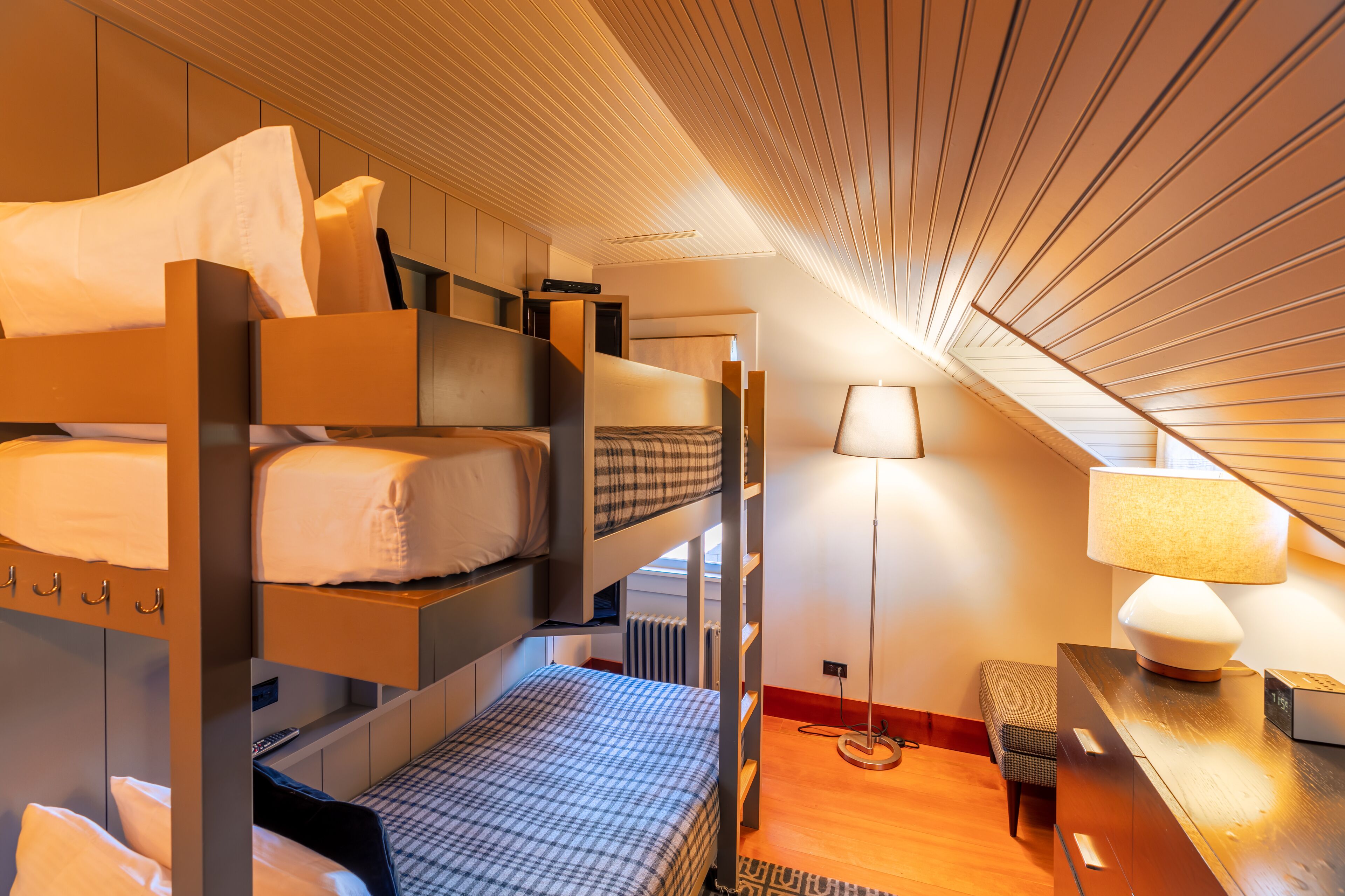 Une chambre avec un lit superposé, un bureau et une lampe, un plancher en bois et un plafond en pente.