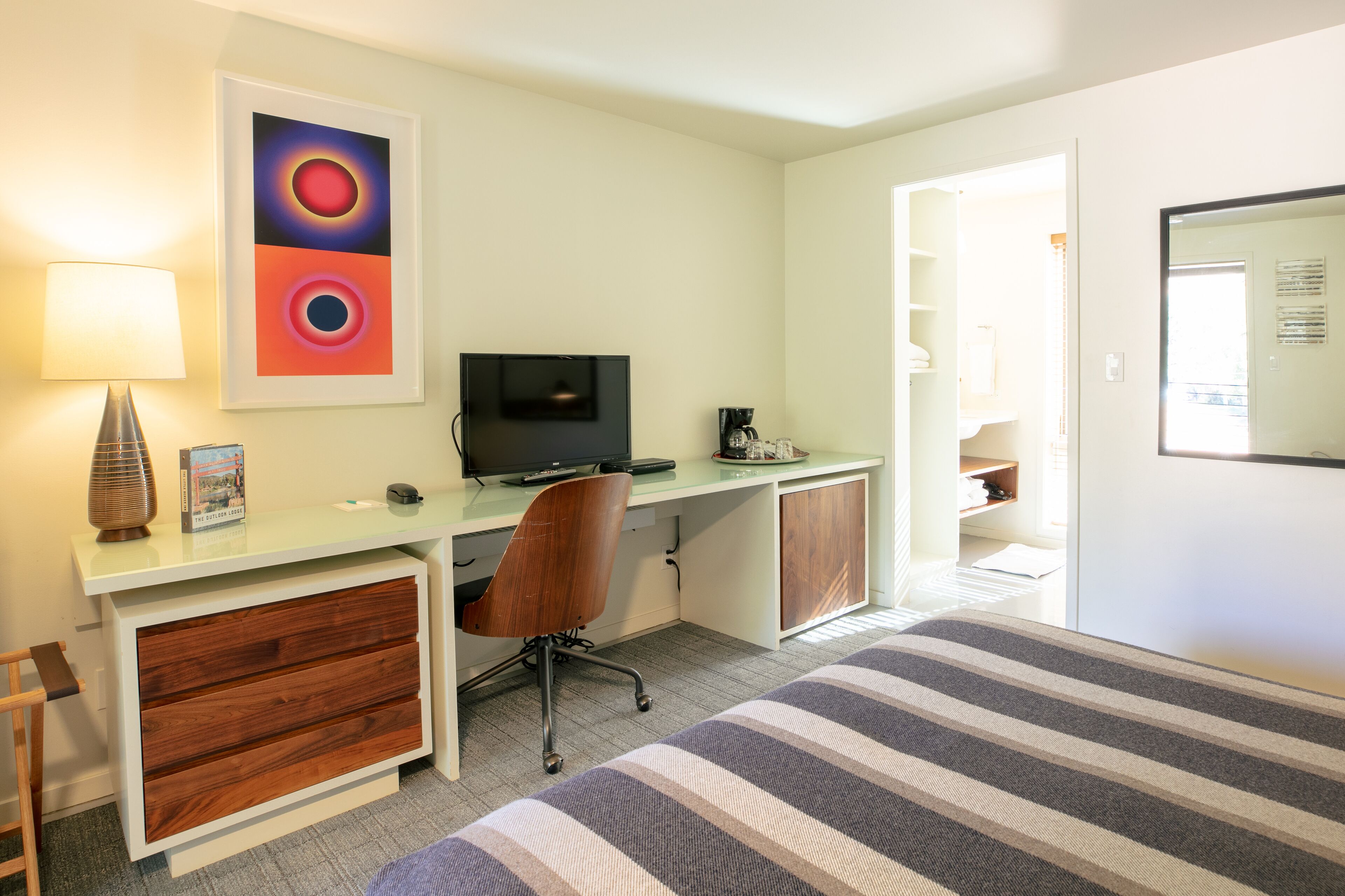 Quarto com cama, escrivaninha com computador, cadeira, luminária e quadro na parede.