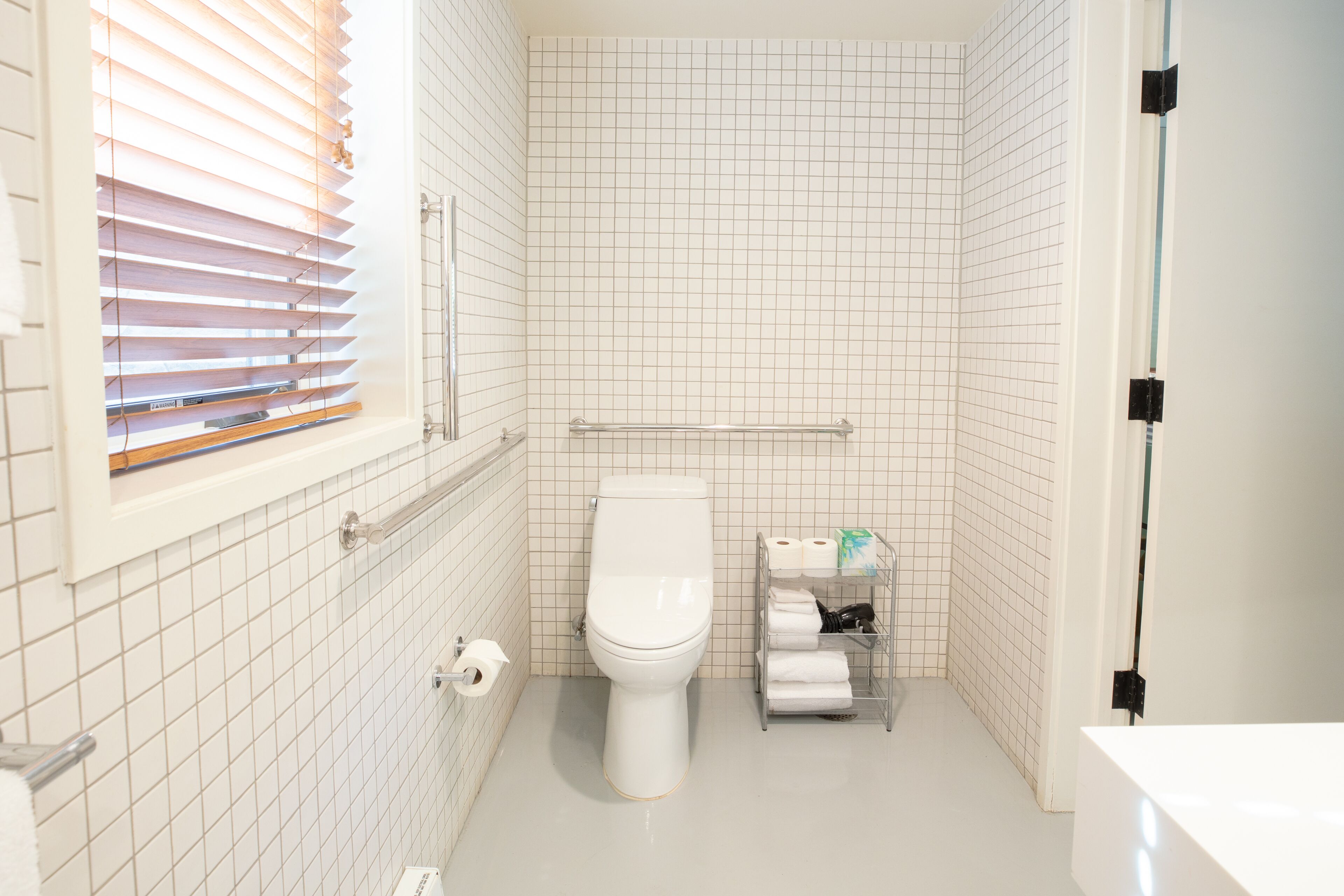 A small, clean bathroom with a toilet, a shelf with towels, and a window with blinds.