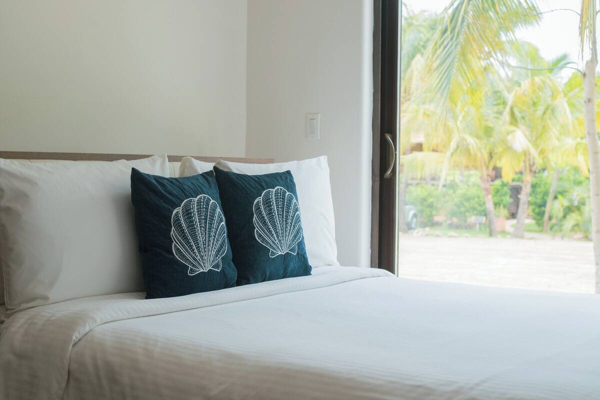 A bed with white and teal pillows, a view of palm trees through a window.