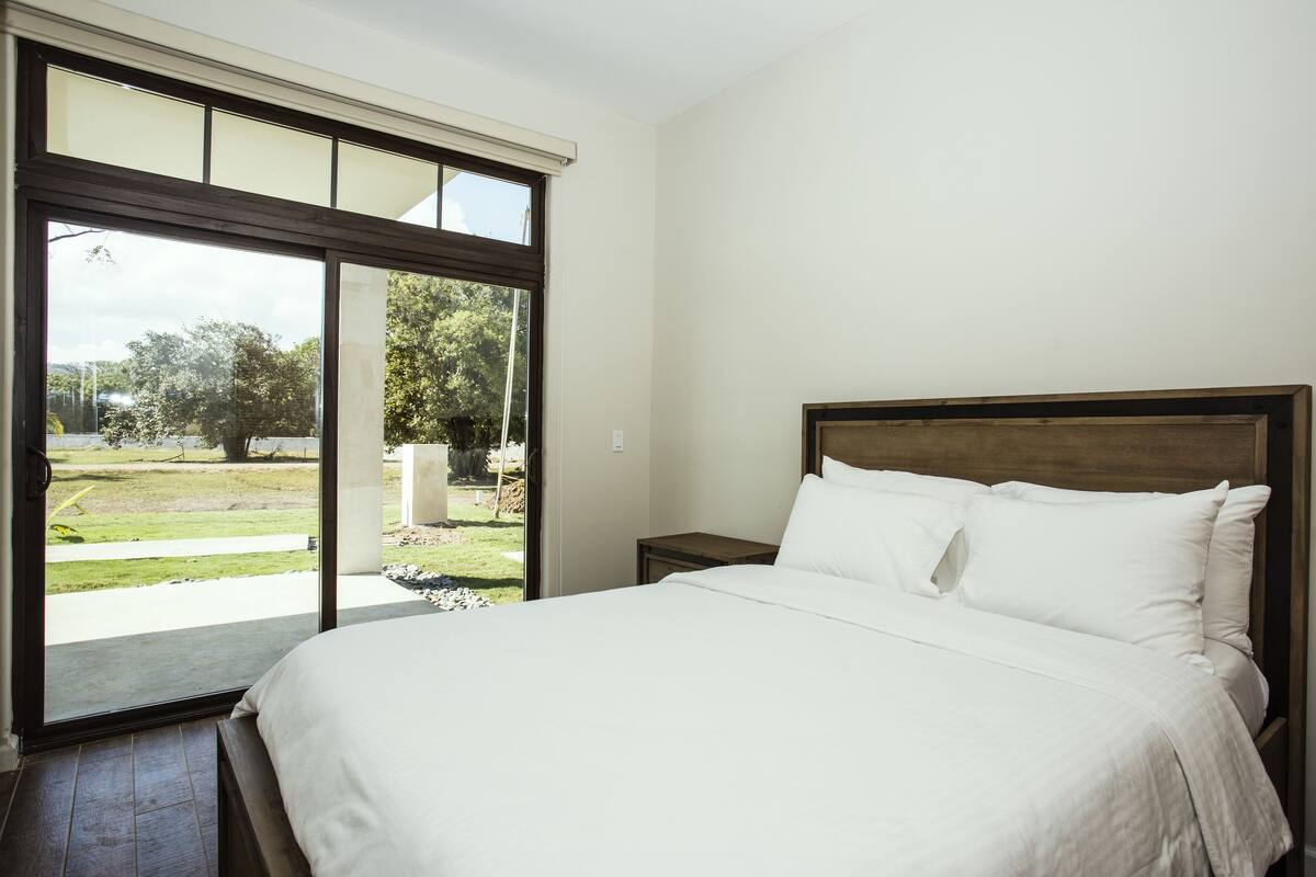 A bedroom with a large bed, a nightstand, and a window with a view of a garden.