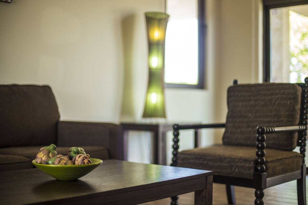 A room with a dark brown sofa, a wooden table with a green bowl, a tall green lamp, and a dark brown chair.