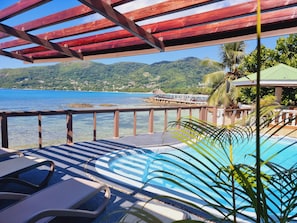 Ein Poolbereich mit Liegestühlen, einer Holzpergola und Blick auf eine Küstenlandschaft mit Bergen und einem Steg.