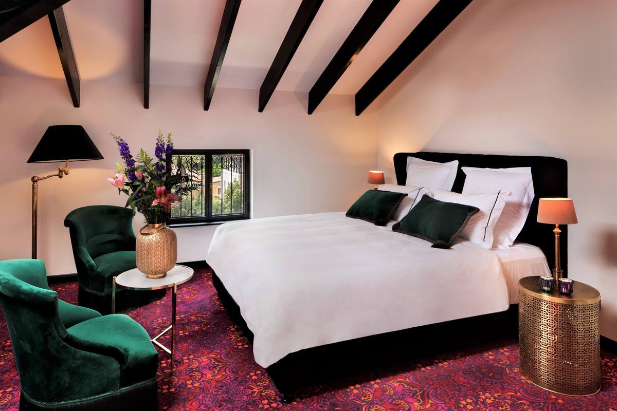 A bedroom with a bed, two chairs, a side table, a lamp, and a window with a view.