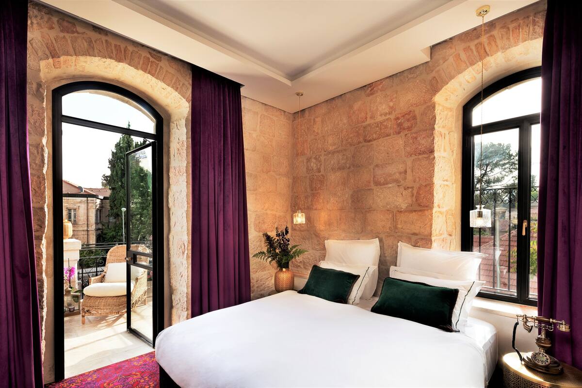 A hotel room with a large bed, two windows with curtains, a small table with a lamp, and a view of the outside balcony.