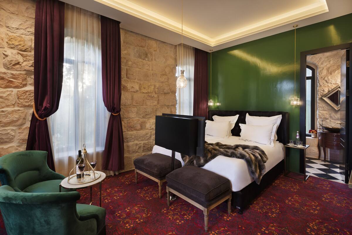 A hotel room with a stone wall, a bed with white linens, a green armchair, and a small table with a bottle and glasses.