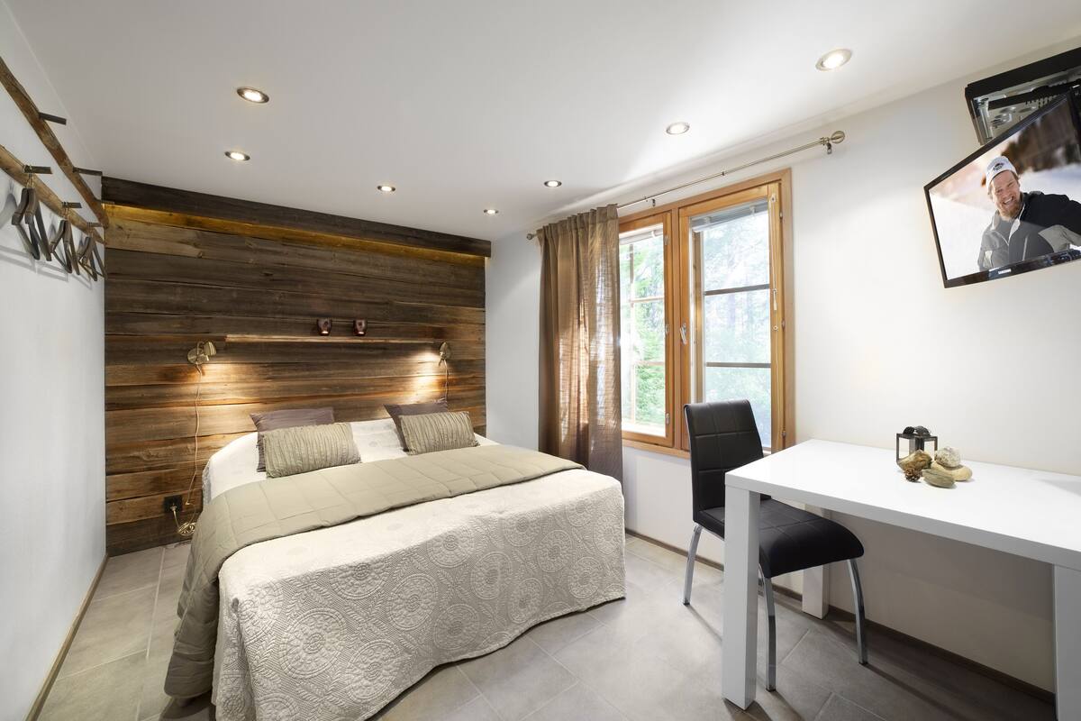 Un dormitorio con cabecera de madera, cama con colcha color beige, escritorio con silla y ventana con cortinas.