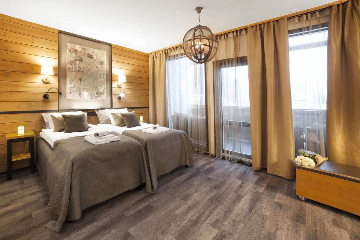 Ein Hotelzimmer mit zwei Betten, Holzwänden, einem großen Fenster mit Vorhängen, einer Kandelaberleuchte und einer hölzernen Bank.