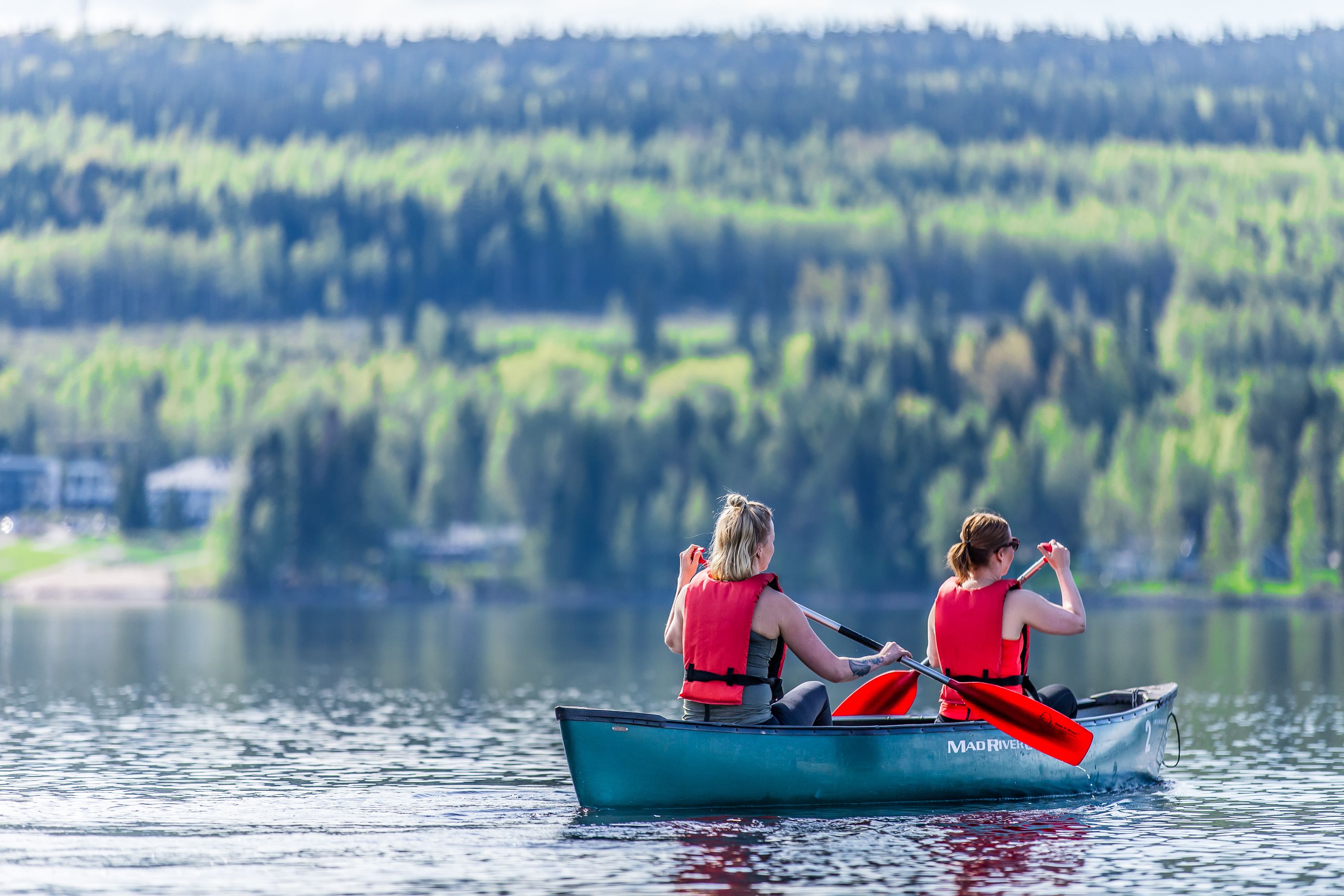 Two people in a small boat on a calm lake with a forested shoreline in the background.