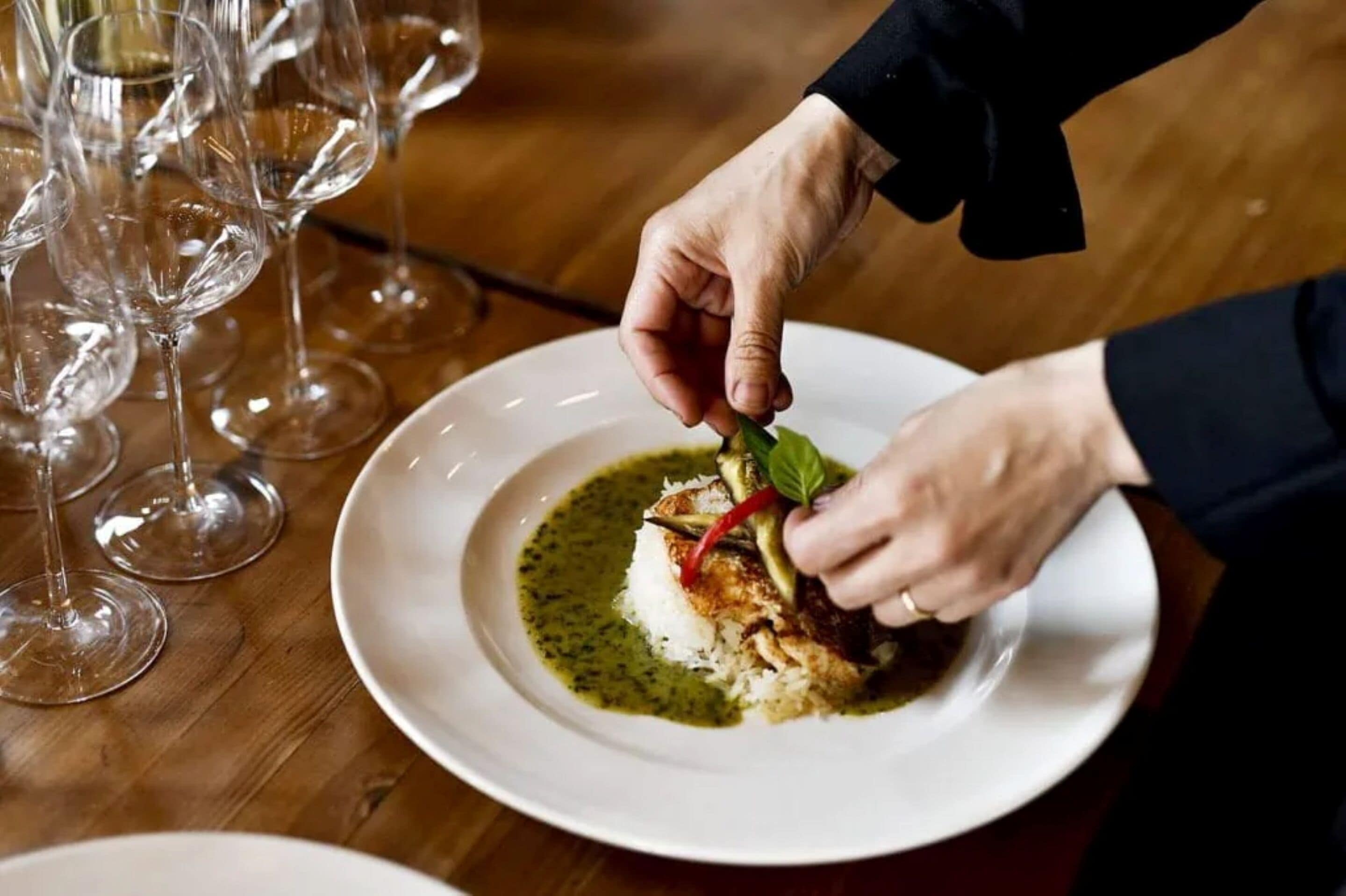 Una persona sirve un plato sobre una mesita de madera, rodeada de copas de vino vacías, con salsa verde.