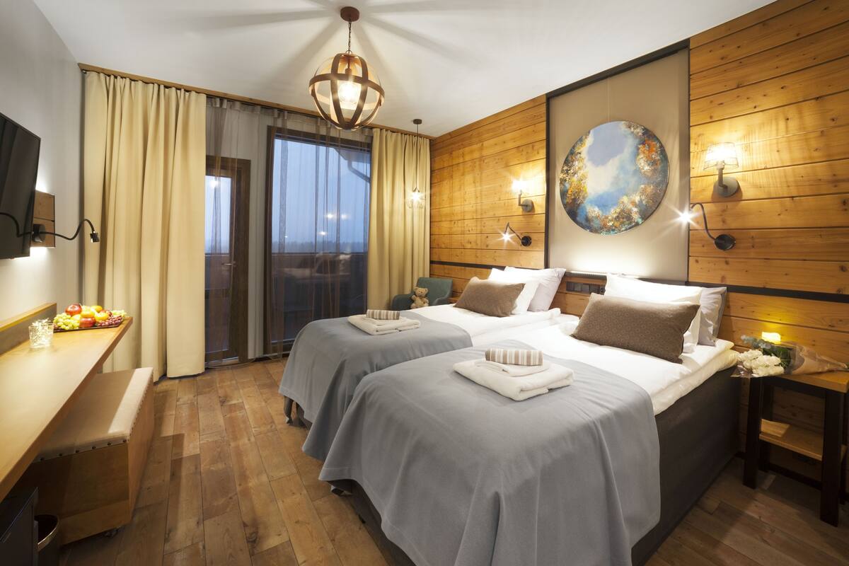 Ein Hotelzimmer mit zwei Betten, Holzvertäfelung, einem großen Fenster mit Vorhängen, einem Esstisch mit Obst und einem an der Wand montierten Fernseher.
