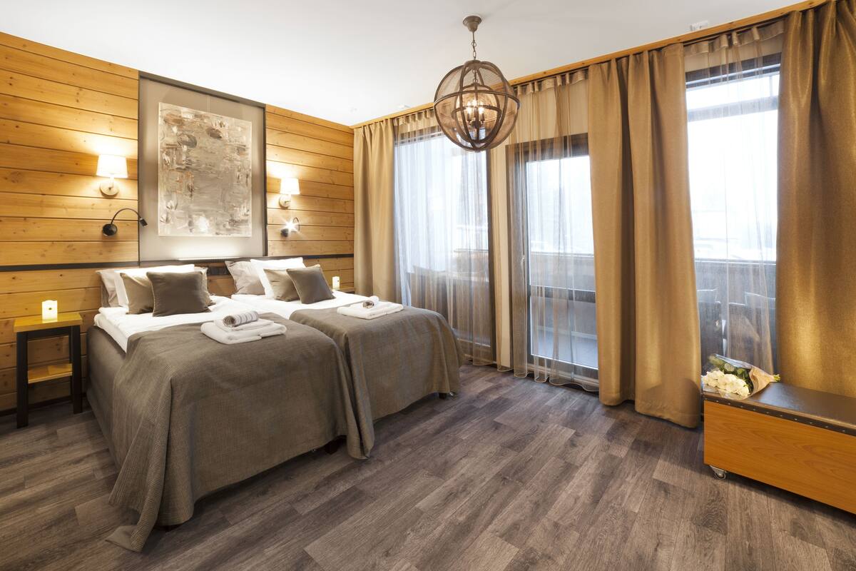 Ein Hotelzimmer mit zwei Betten, Holzvertäfelung, einem großen Fenster mit Vorhängen, einer Kristallleuchtre und einem kleinen Tisch mit einer Lampe.
