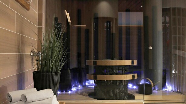 Un espacio moderno de spa con un banco de madera, una zona de estar con toallas, un reloj decorativo grande y una partición de vidrio.