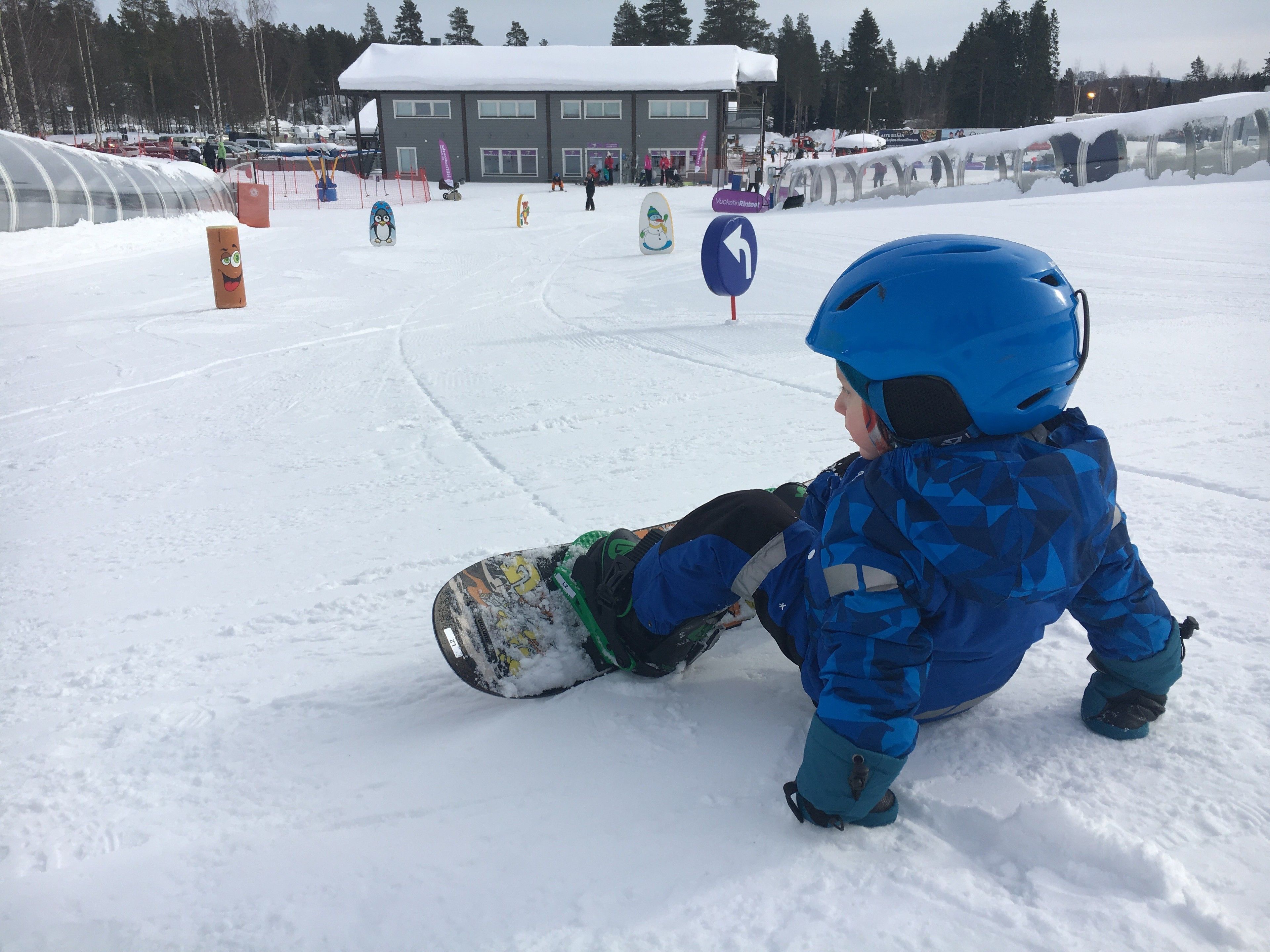 Un niño con equipo de invierno azul y una tabla de snowboard, sentado sobre la nieve.