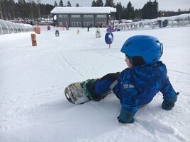 Un niño con equipo de invierno azul y una tabla de snowboard, sentado sobre la nieve.