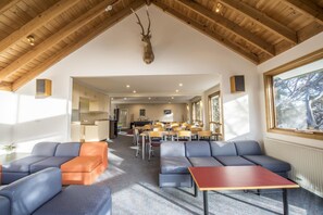 A spacious room with a high ceiling, wooden beams, a mounted deer head, blue sofas, a red table, and large windows offering a view of trees. - Terama Ski Lodge (Mount Buller)