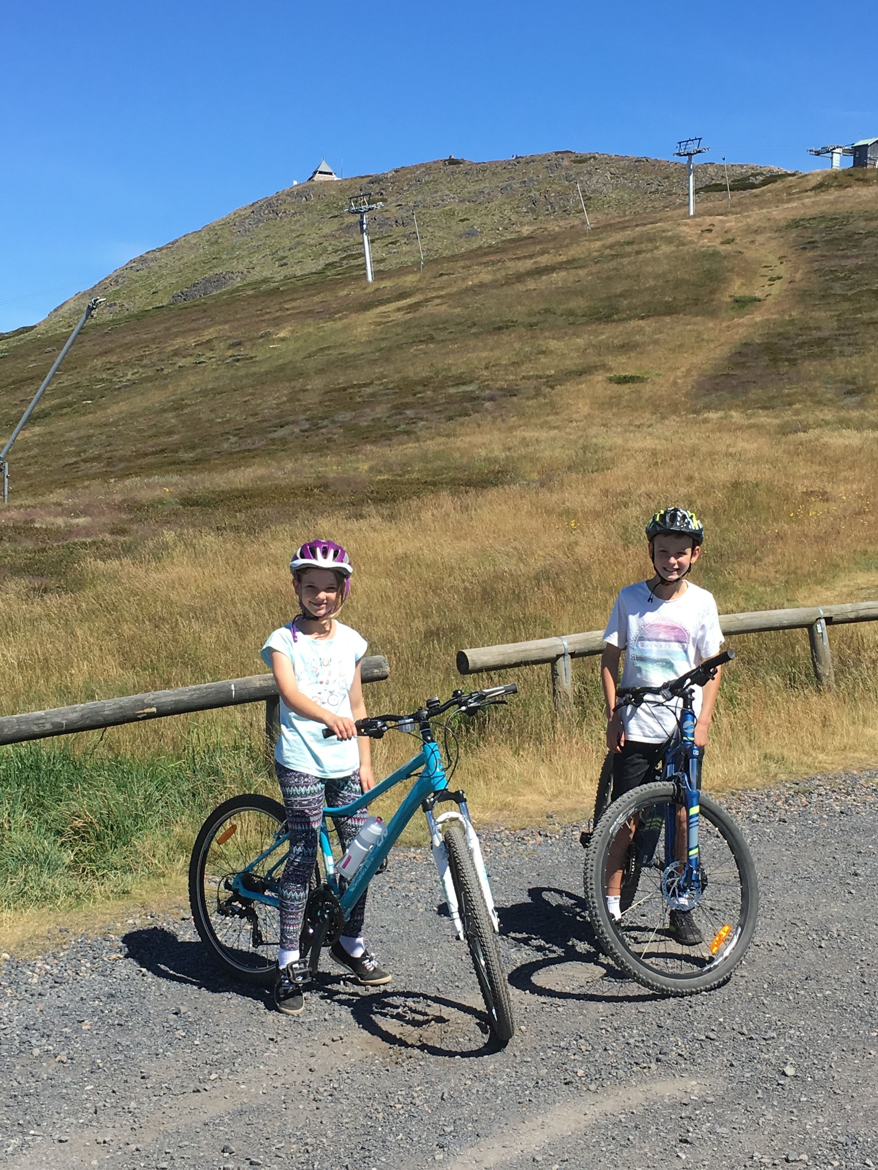 Dos niños de pie junto a bicicletas en un sendero de grava con una valla de madera y un paisaje montañoso al fondo.