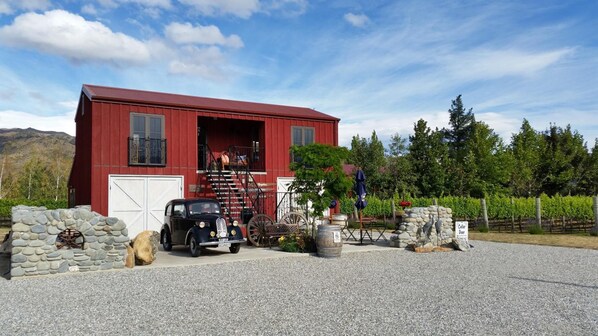 A red barn with a white garage door, a vintage car, and a stone wall with a large tree in front.
