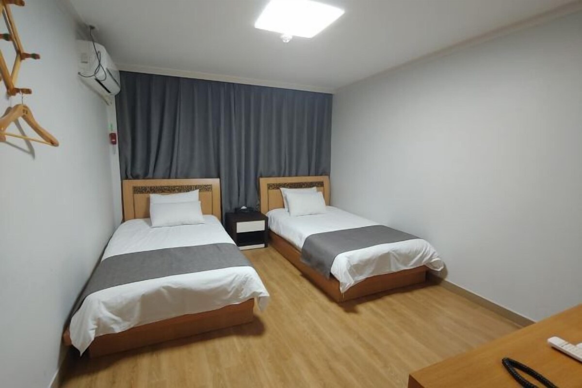 A hotel room with two single beds, a wooden floor, a desk with a chair, and a window with curtains.