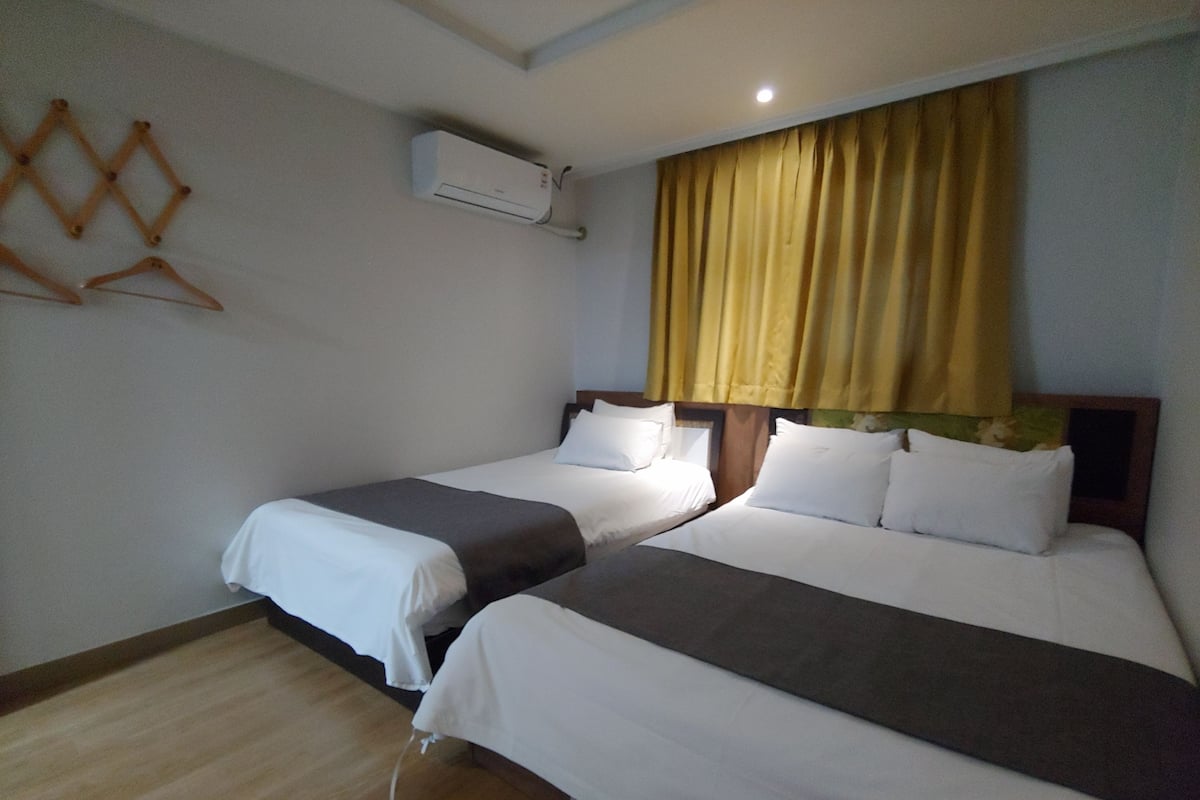 A hotel room with two beds, a wooden floor, a wall-mounted air conditioner, and a window with yellow curtains.