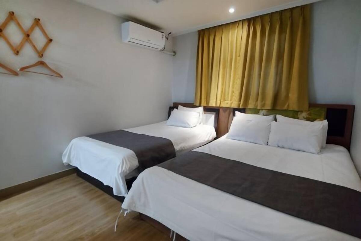 A hotel room with two beds, a wooden floor, a wall-mounted air conditioner, and a window with yellow curtains.