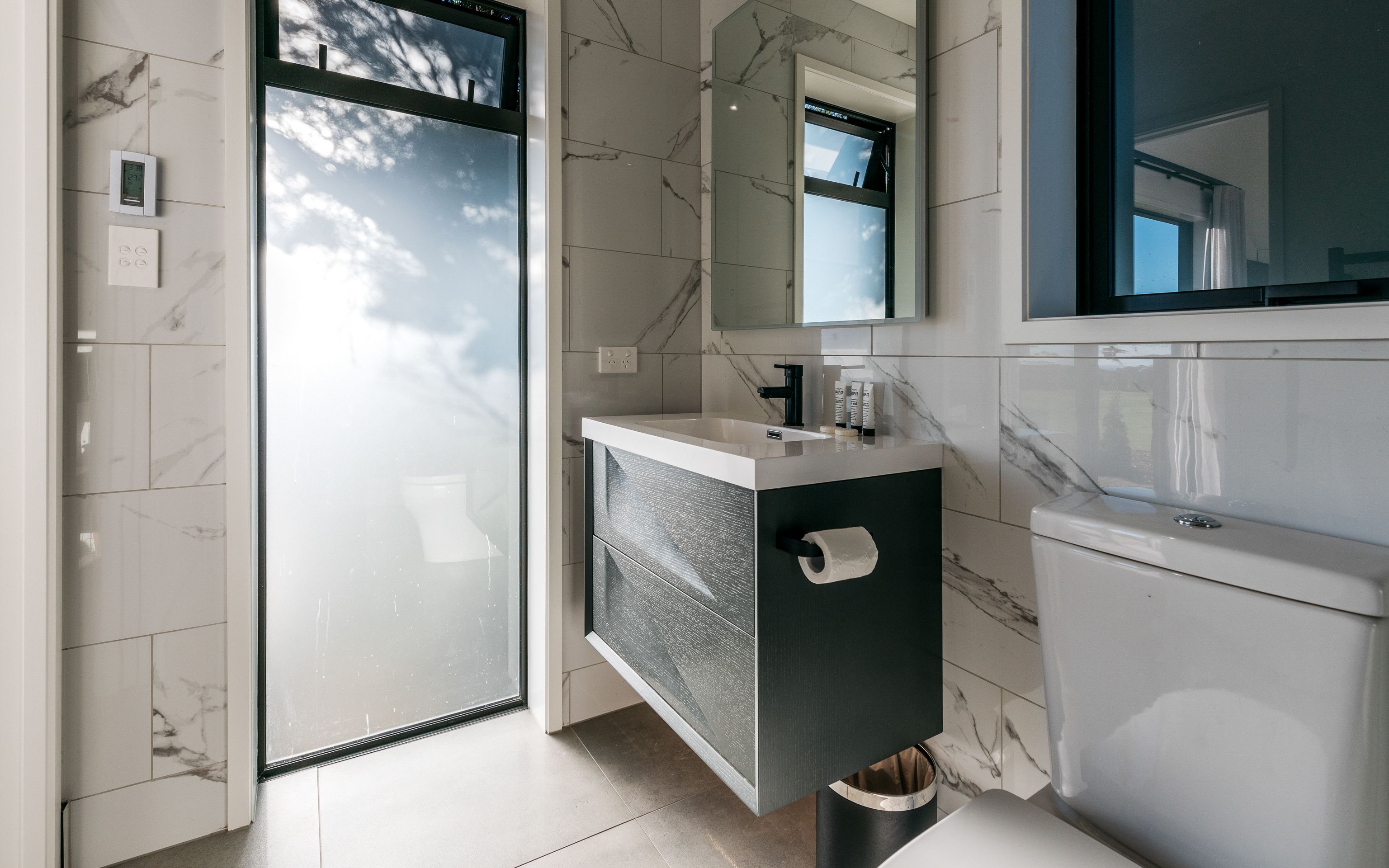 A modern bathroom with a glass shower door, a marble-tiled wall, a white sink with a dark cabinet, and a large mirror.