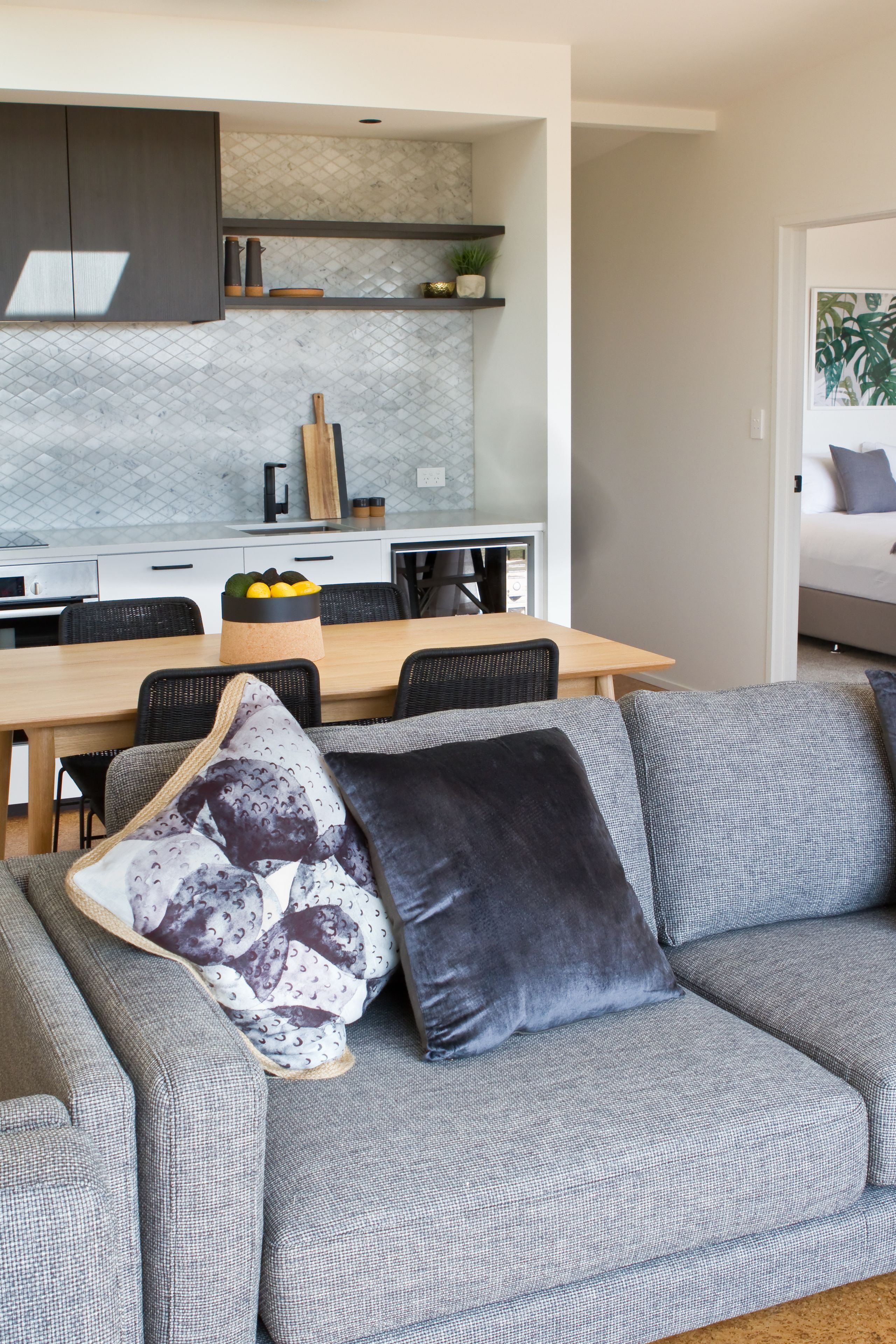 A modern living room with a grey sofa, a wooden dining table, and a kitchen in the background.