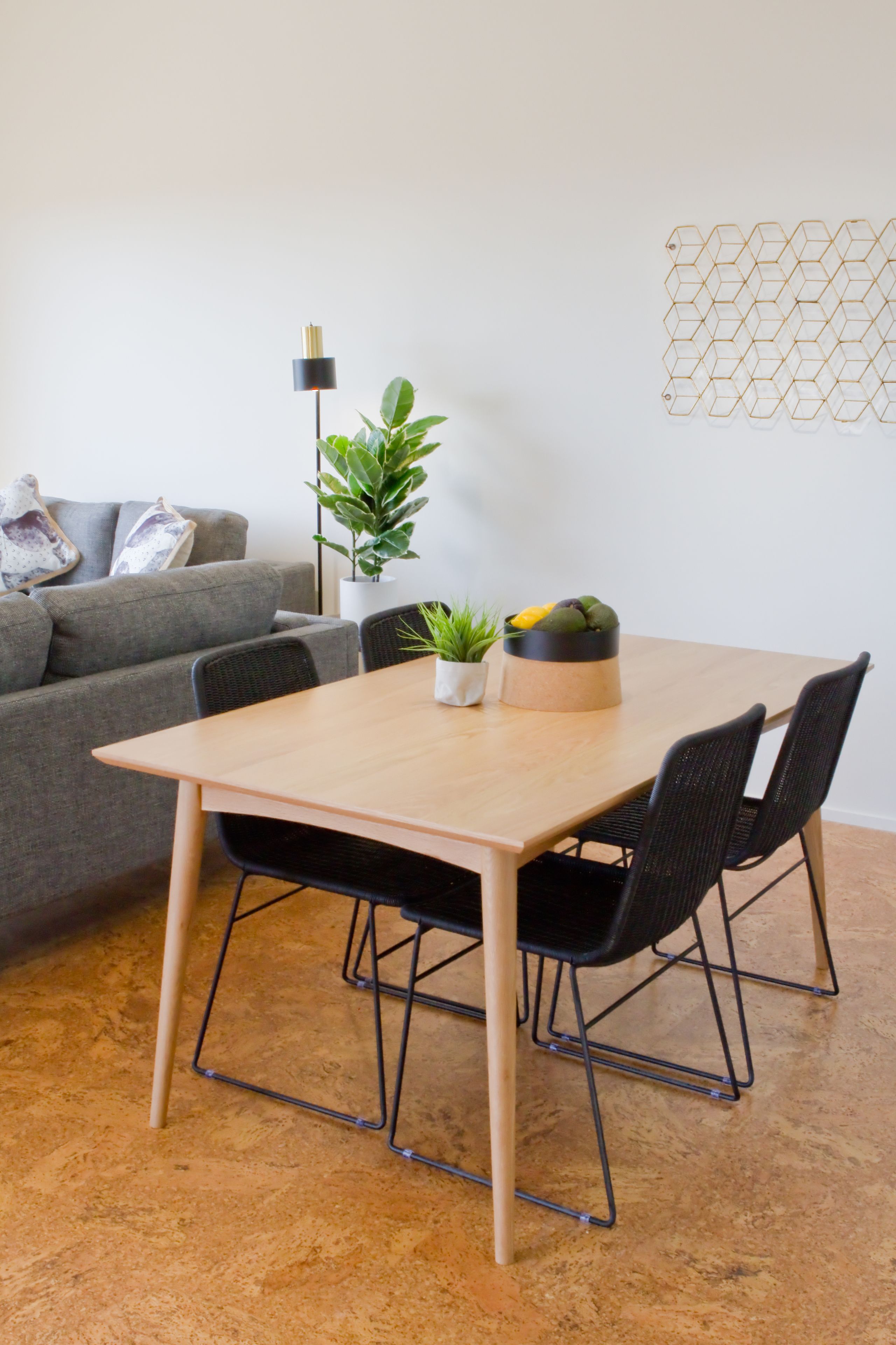 A dining area with a wooden table, black chairs, a grey sofa, a floor lamp, and a decorative wall piece.