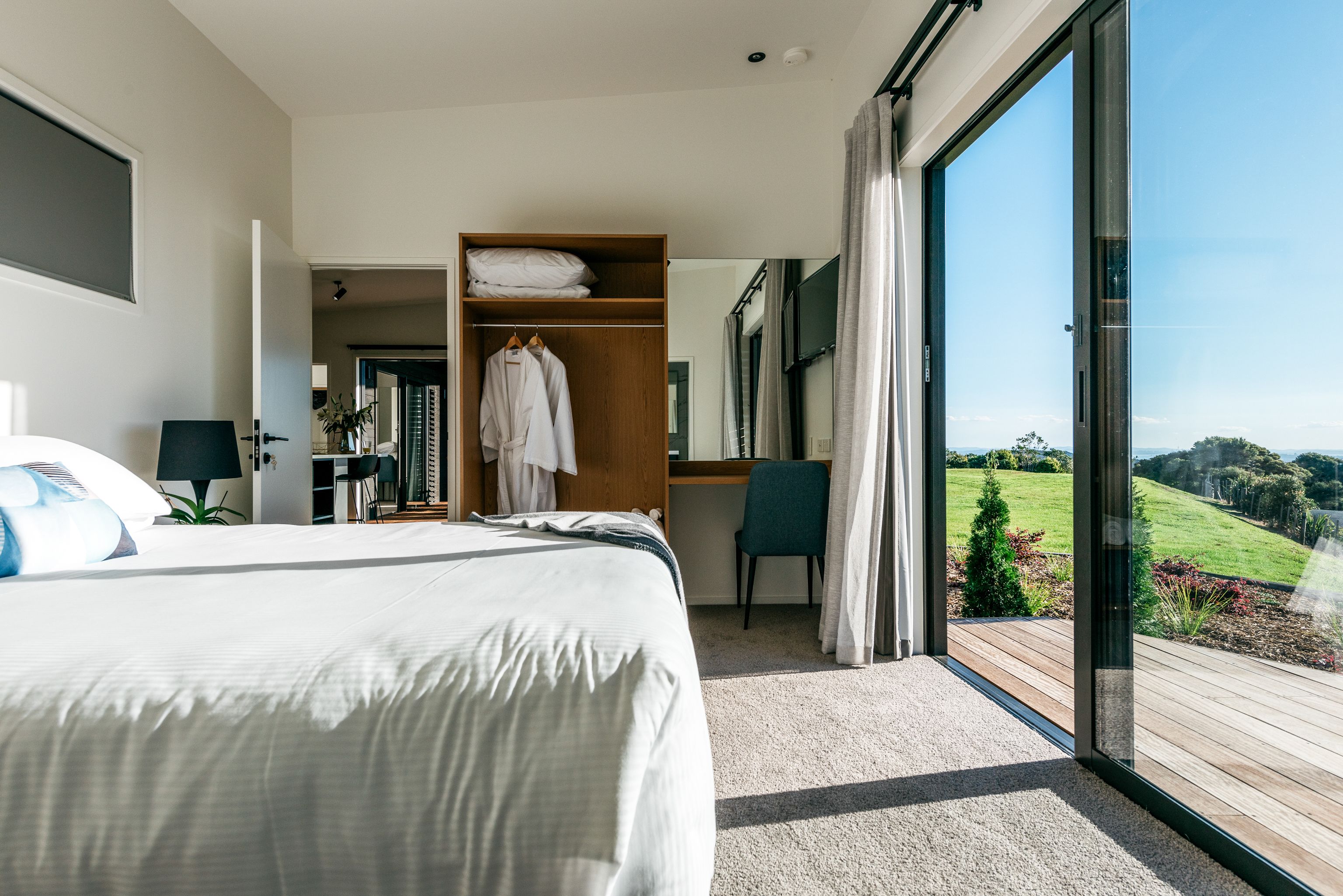 A modern hotel room with a large bed, a wooden wardrobe, a desk, and a view of the outdoors.