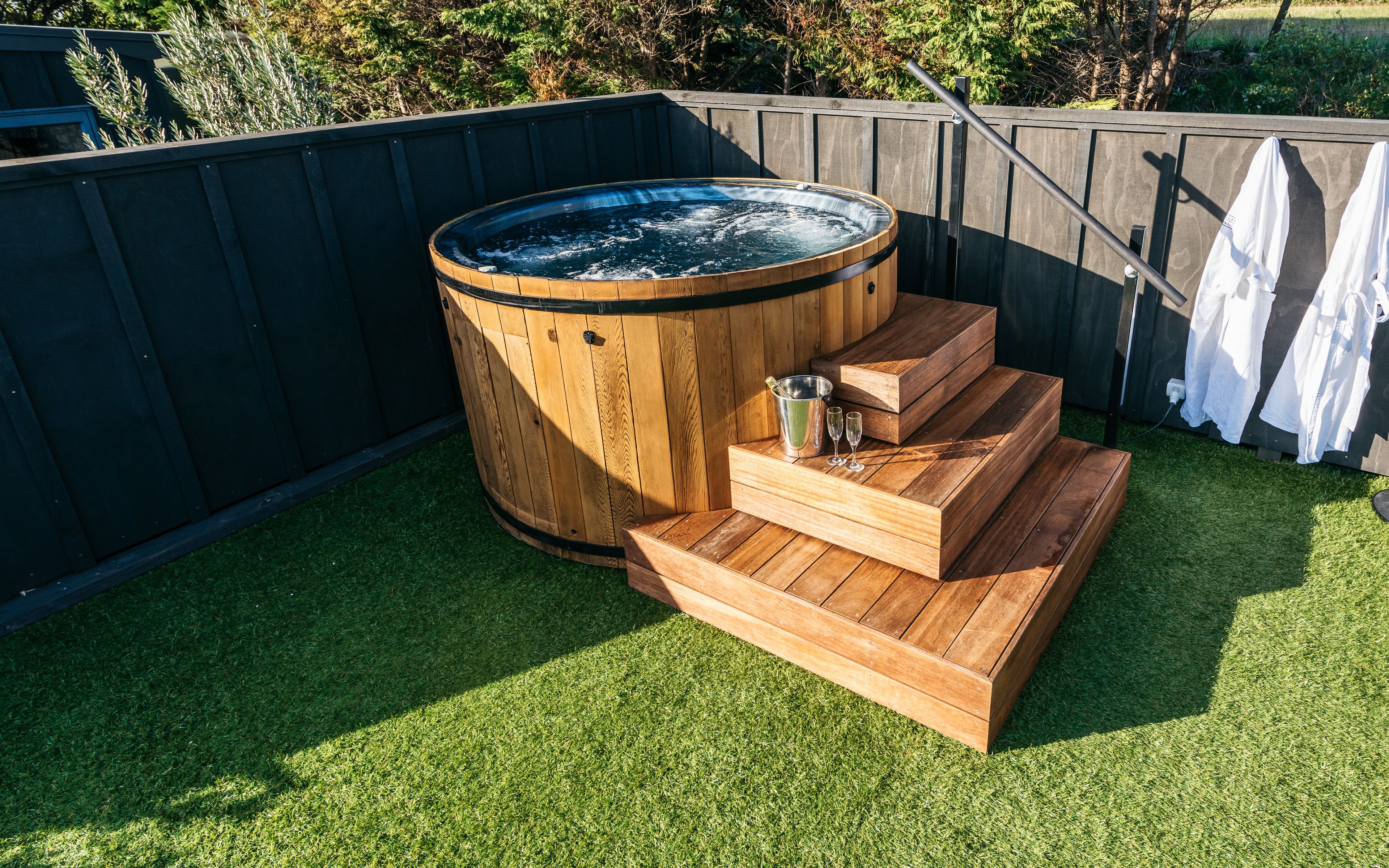 Un jacuzzi con escalones de madera y un cubo metálico, rodeado por una valla de madera y césped artificial verde.