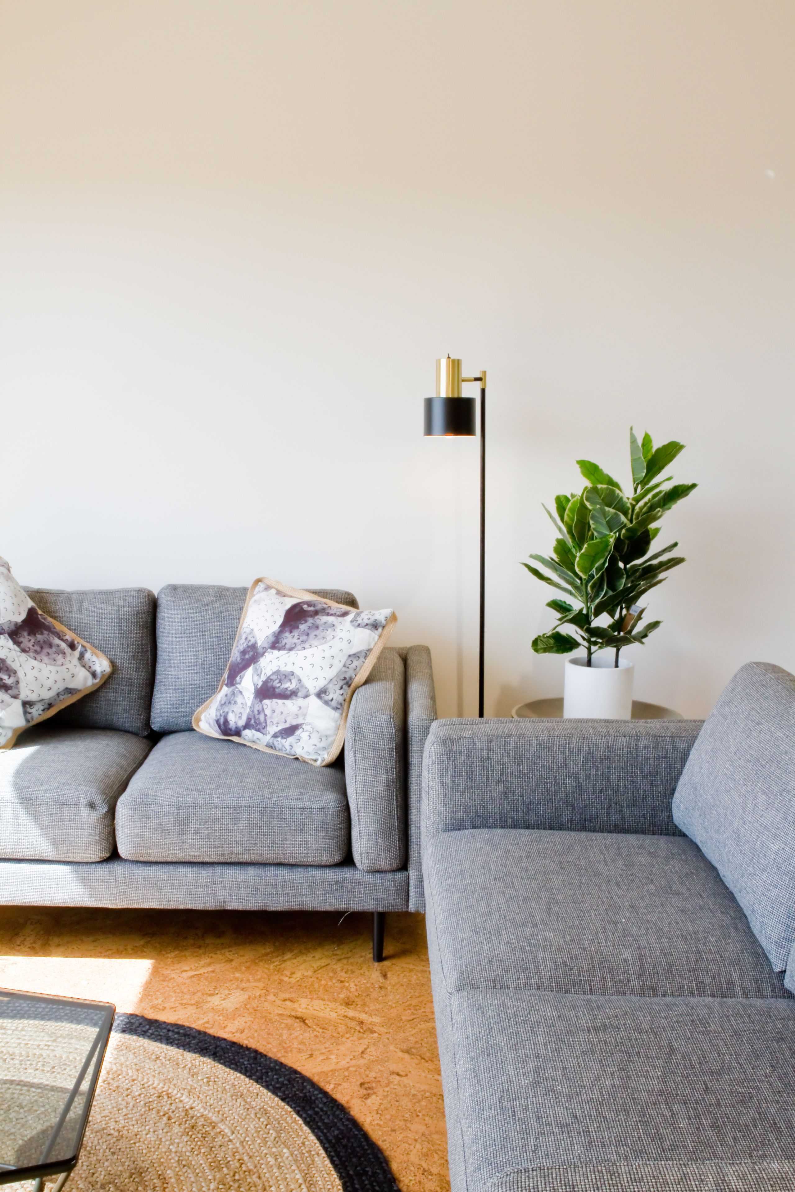 A modern living room with a grey sofa, patterned cushions, a small side table, a floor lamp, and a potted plant.